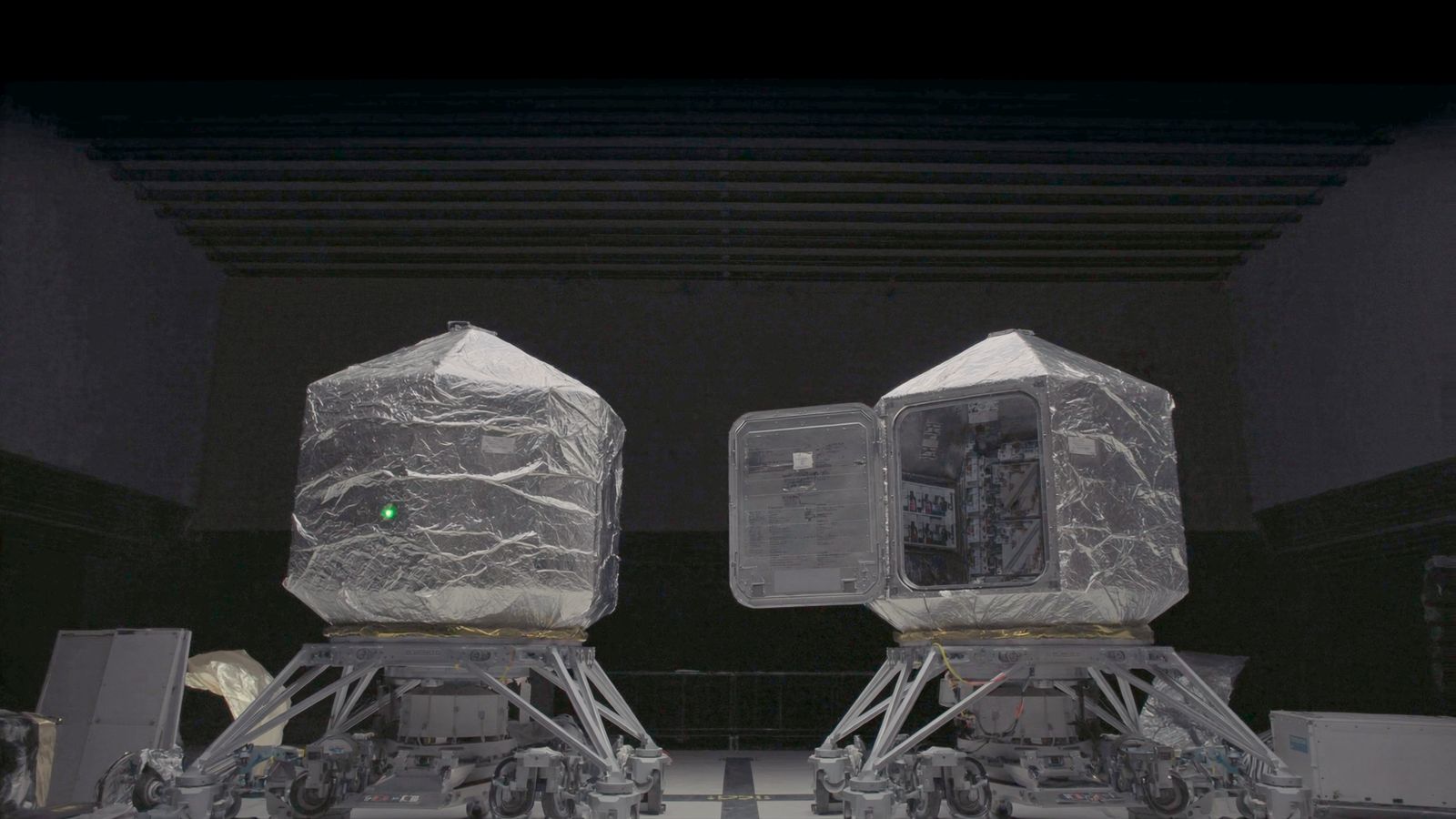 Two identical satellite buses wrapped in silver thermal blanket on integration stands in a darkened assembly hall, the right-hand one with an access panel swung open on its equipment shelves, a single chartreuse status light on the left-hand bus