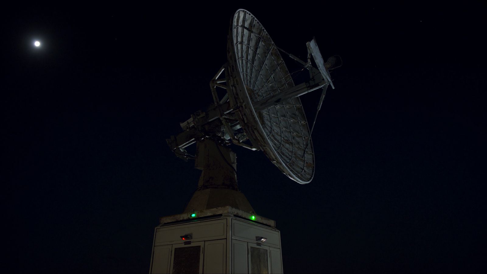 A 13-metre TT&C tracking dish on a concrete pedestal on a dark headland at night, one chartreuse status light on the pedestal housing
