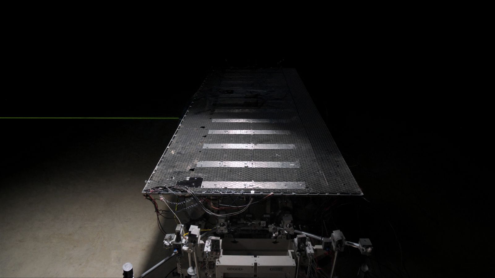 A satellite structural panel and harness on an assembly stand in a dark cleanroom, one chartreuse alignment laser crossing the frame