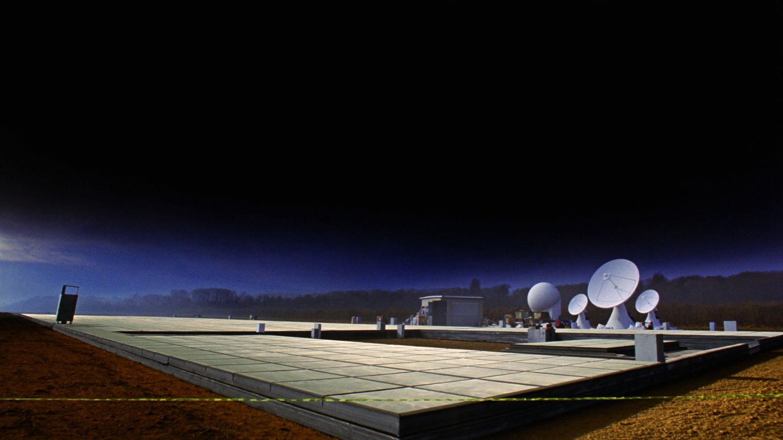 A newly built gateway earth station before sunrise, parabolic antennas and a site cabin beyond a run of fresh concrete pads, with a thin chartreuse survey line across the foreground
